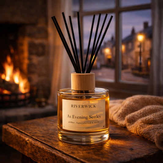 Riverwick As Evening Settles reed diffuser glowing beside a fire while evening falls outside a window overlooking a softly lit village street.